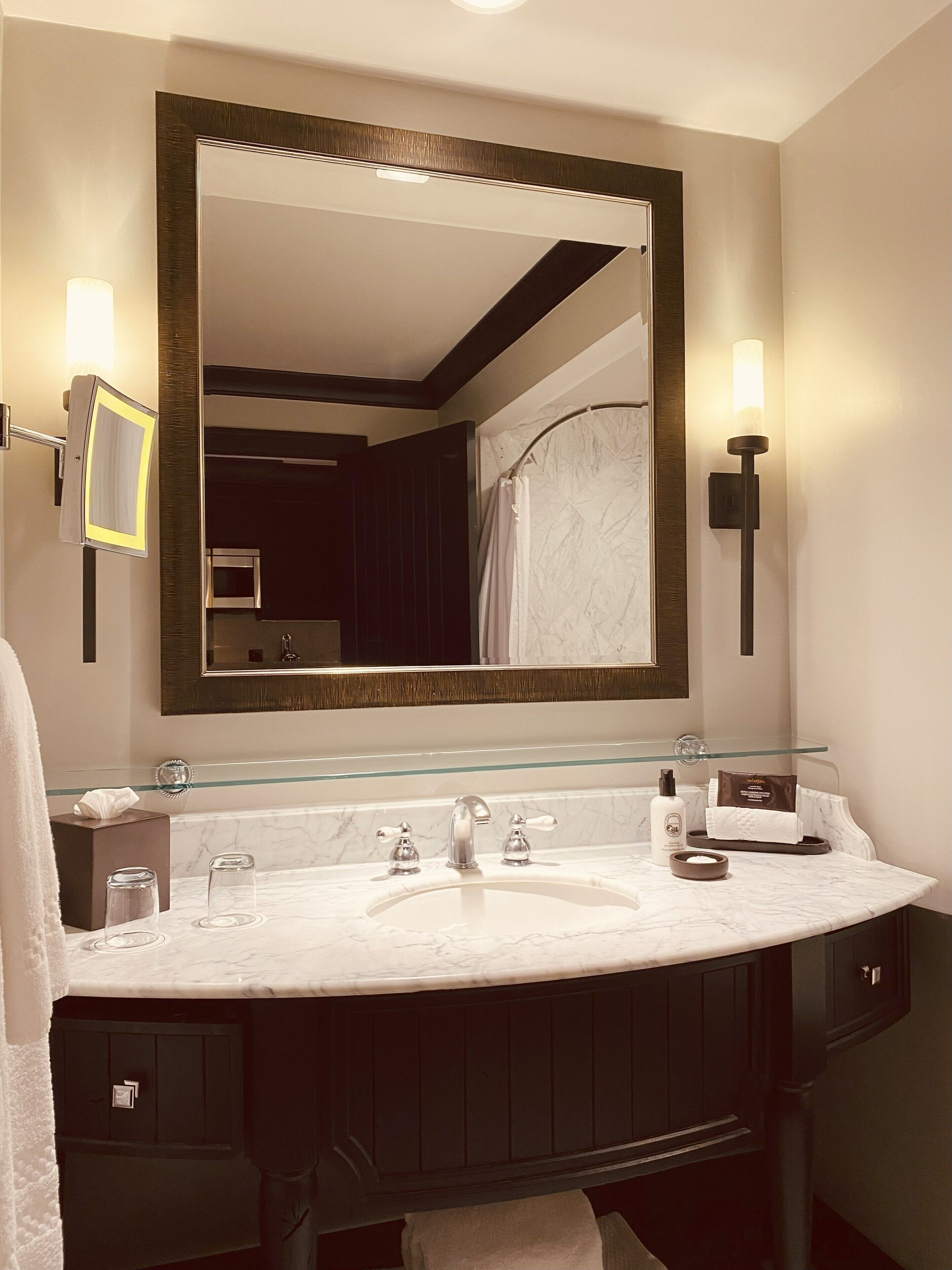 A bathroom with a large mirror, a marble sink, two wall-mounted lights, and a dark wooden vanity.
