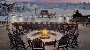 Ein mit Schnee bedeckter Außenbereich mit Adirondack-Stühlen, die um eine zentrale Feuerstelle angeordnet sind, mit Blick auf einen Skiort mit Gebäuden und einem Sessellift.