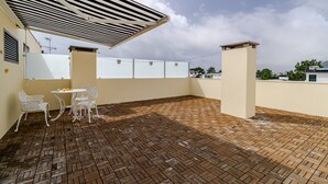 Um terraço no telhado com piso de tijolos, cadeiras de metal branco, uma pequena mesa redonda e um toldo listrado.