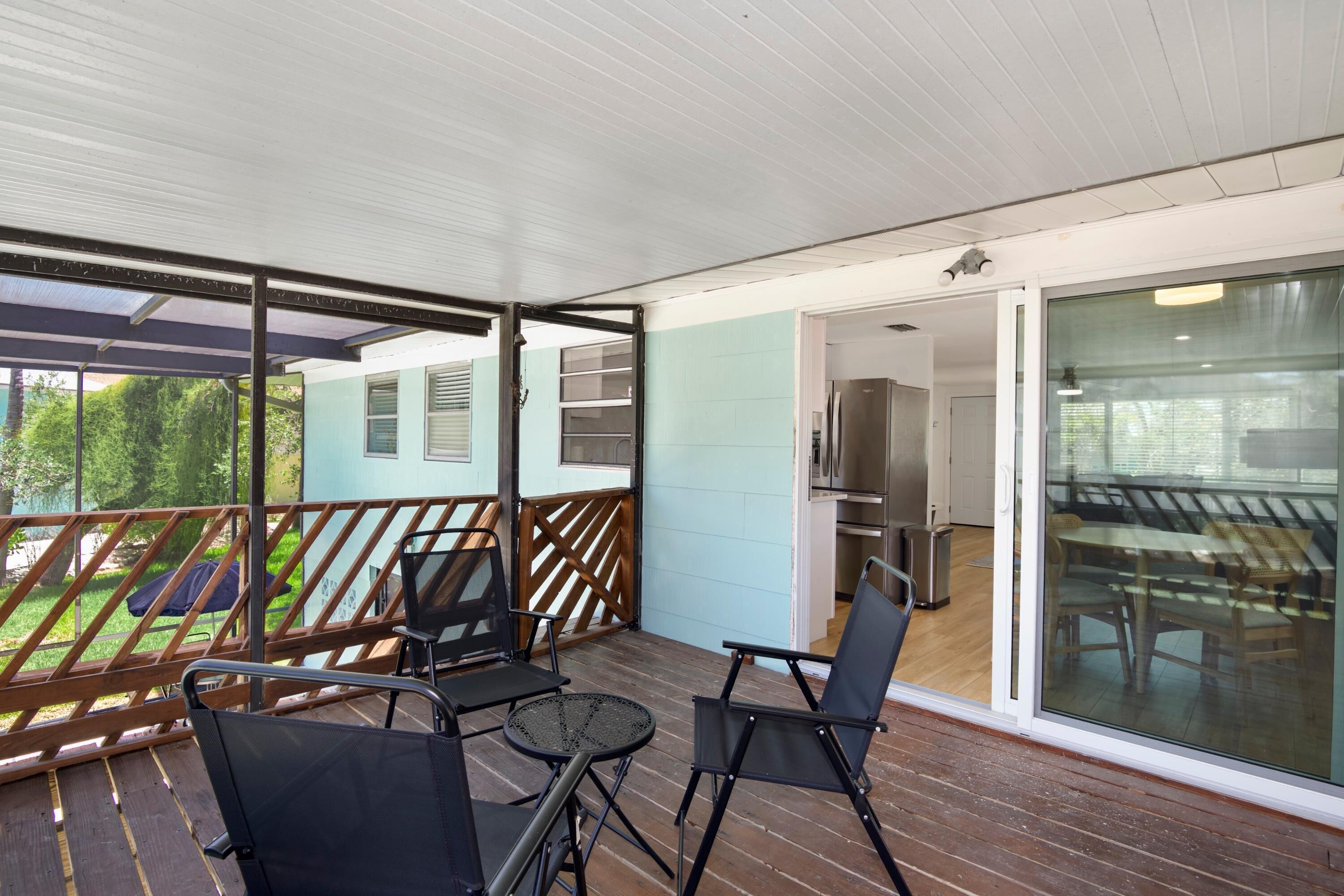 A covered outdoor area with a wooden deck, metal chairs, and a glass door leading to an interior space with a kitchen and dining area.
