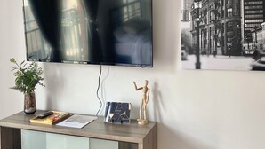 A modern living room with a flat-screen TV mounted on the wall, a wooden console table with a vase and remote control, and a framed picture of a cityscape. - Studio urbain à Ville-Marie – 309, emplacement central à Montréal (Montréal)