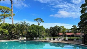 A clear swimming pool with lounge chairs and a fence, surrounded by trees and a building with a red roof.