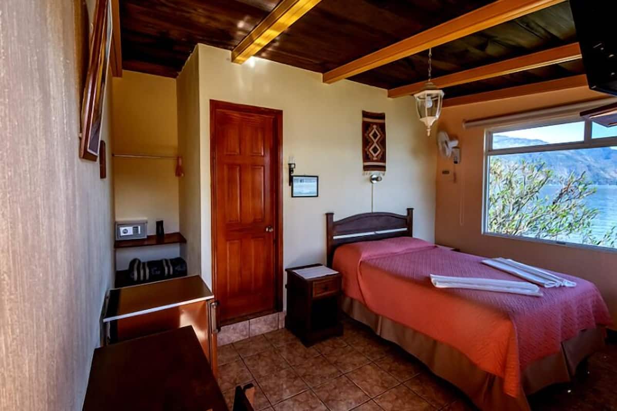 A bedroom with a bed, a nightstand, a wooden door, a window with a view, and a small desk with a chair.