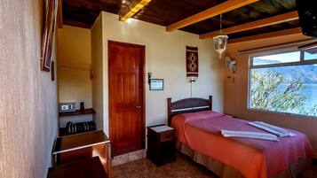 A bedroom with a bed, a nightstand, a wooden door, a window with a view, and a small desk with a chair.