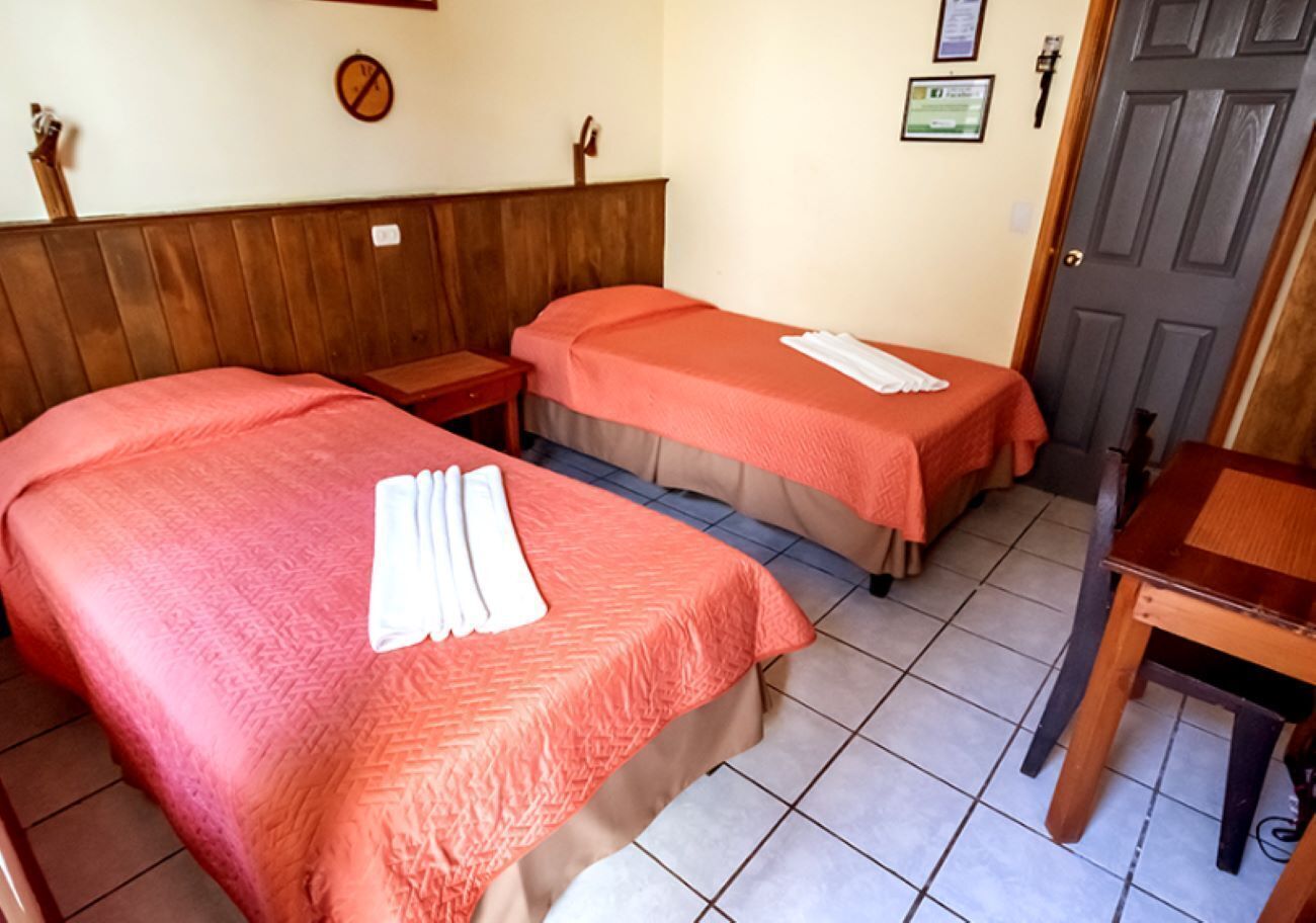 A room with two beds, each with a red bedspread and white towels. There is a wooden headboard, a small wooden table, and a door in the background.