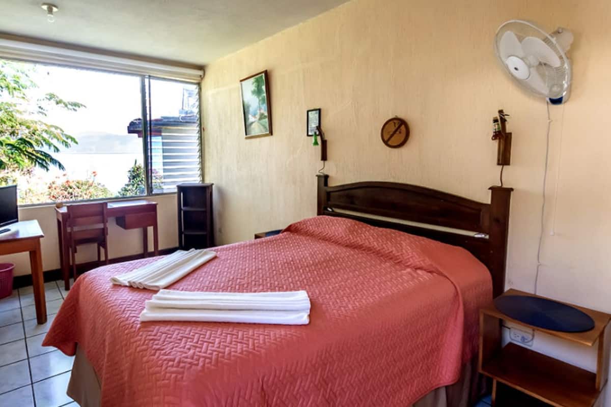 A hotel room with a bed, a desk, a chair, a fan, and a window with a view.