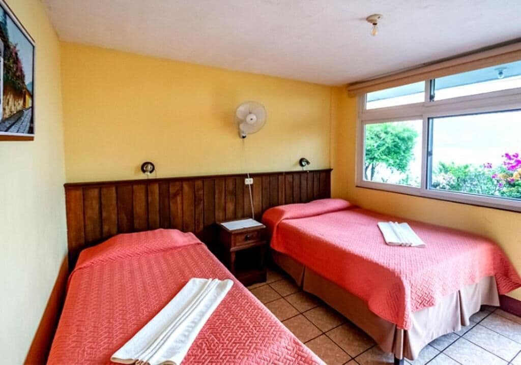 Una habitación con dos camas, un cabecero de madera, un ventilador y una ventana con vistas a zonas verdes.