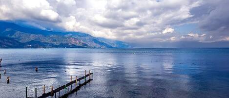 A calm lake with a wooden dock extending into the water, surrounded by mountains and a partly cloudy sky.