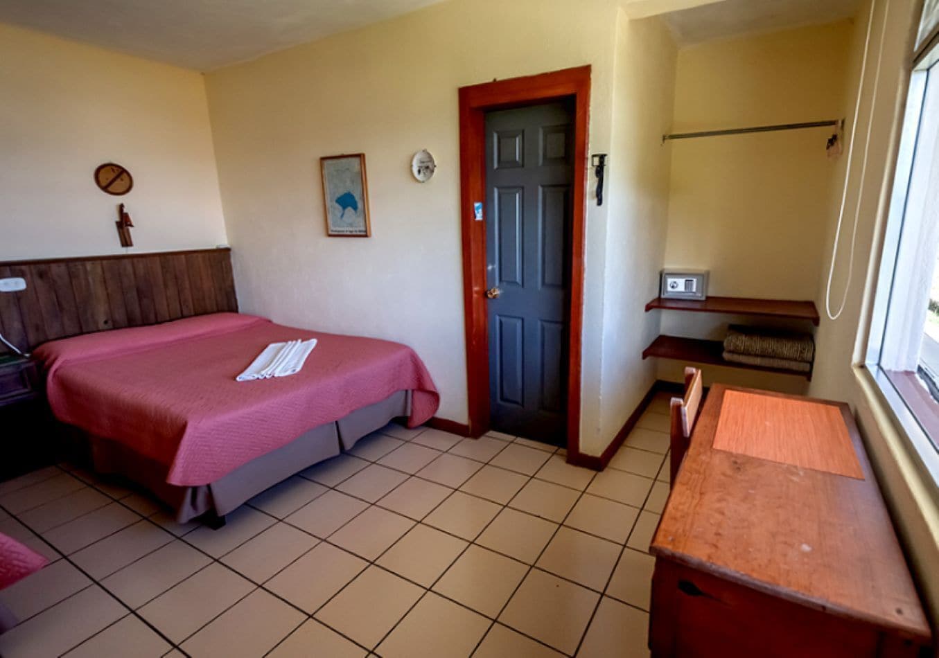 A small, simple room with a bed, a desk, a chair, a door, and a window.