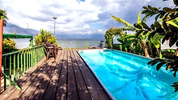 A wooden deck with a pool, lounge chairs, and a view of the sea. - Hotel Sak'cari El Amanecer (San Pedro La Laguna)
