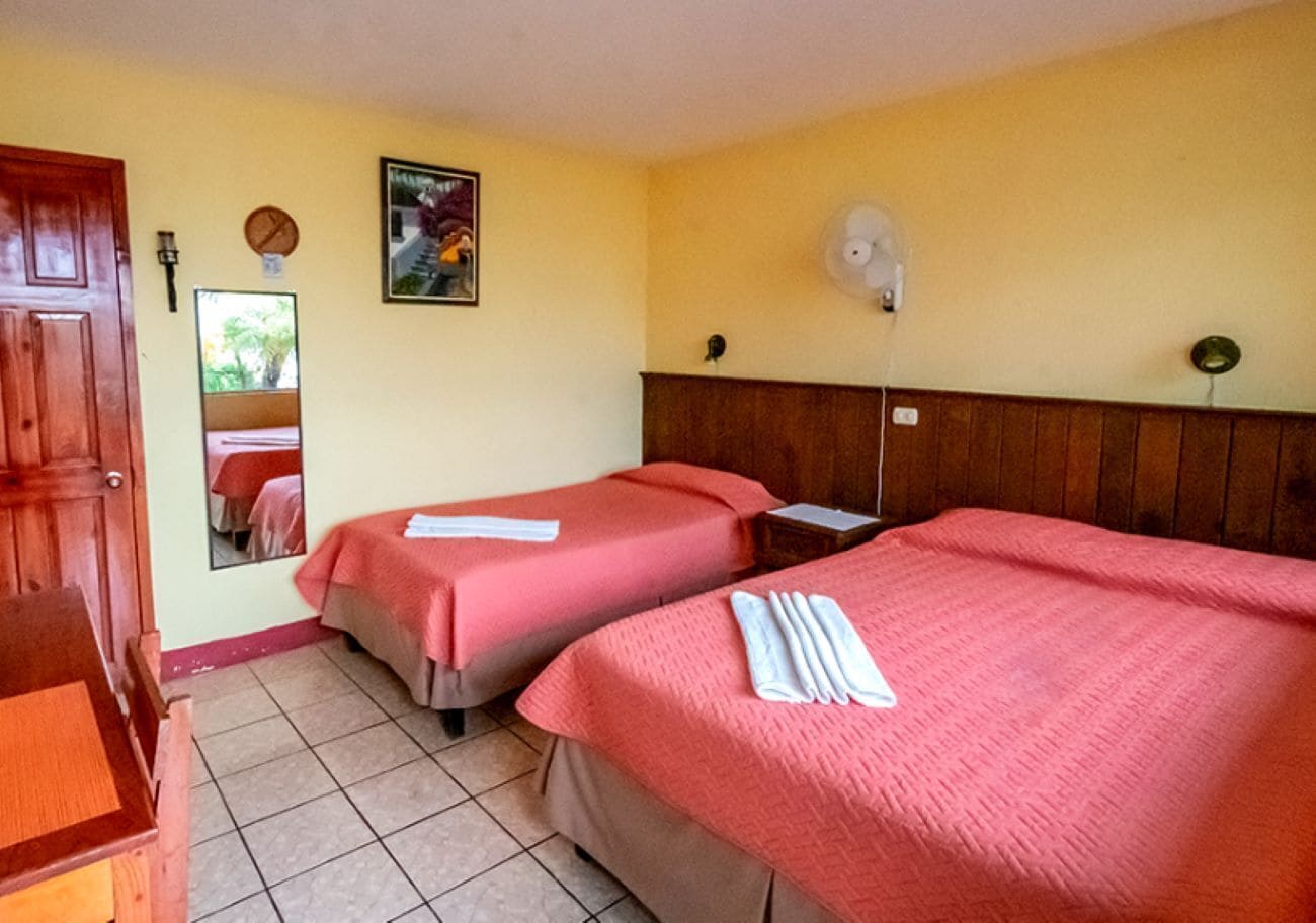 A hotel room with two beds, a wooden door, a mirror, and a wall-mounted fan.