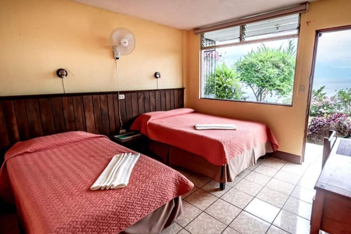 A room with two beds, each with a red bedspread and a white towel on the bed. There is a window with a view of greenery and a body of water.