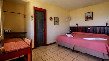 A hotel room with a bed, a desk, a chair, a door, and a wall-mounted fan.