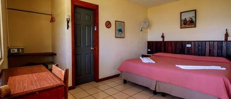 A hotel room with a bed, a desk, a chair, a door, and a wall-mounted fan.