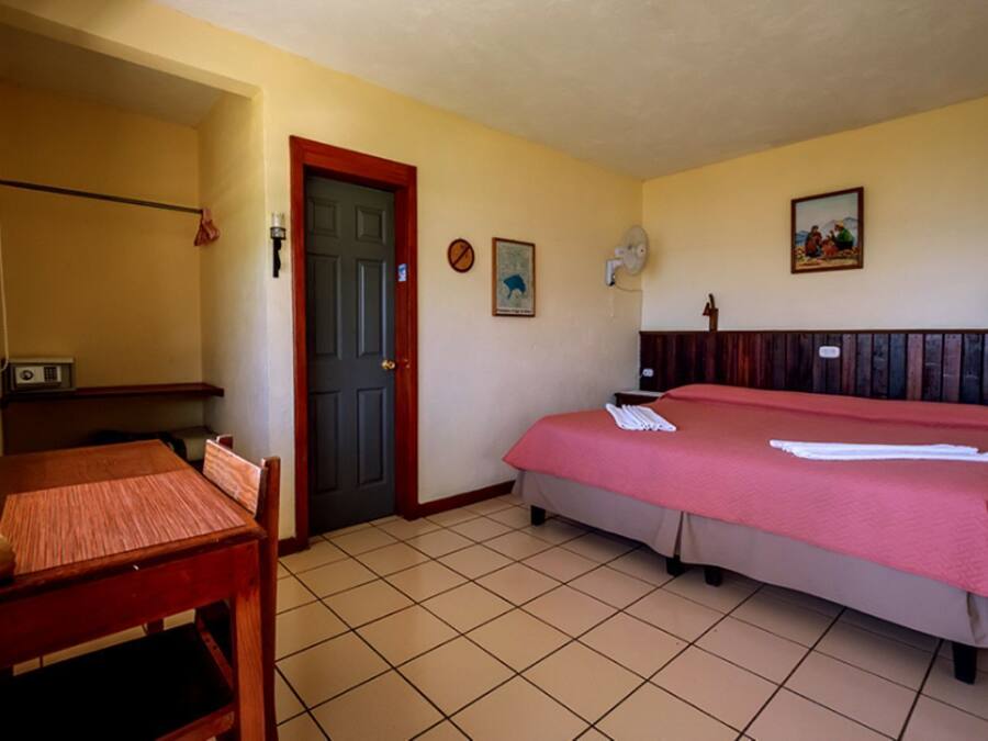 A hotel room with a bed, a desk, a chair, a door, and a wall-mounted fan.