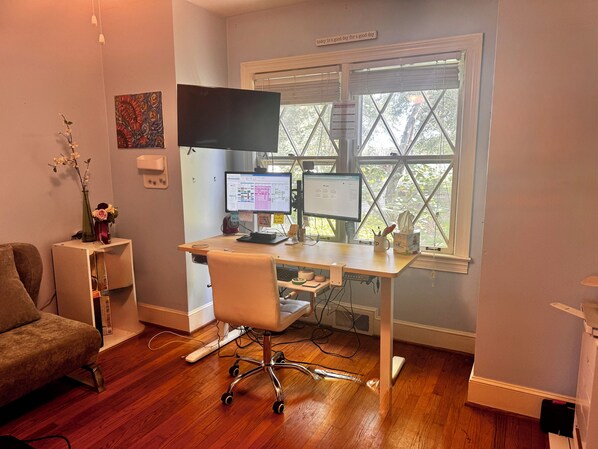 A home office with a desk, two monitors, a chair, a couch, and a window with blinds.