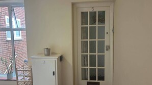 A white door with a frosted glass panel, a small potted plant on a white cabinet, and a wooden rack with a doormat in front. - Homely 2-bed Apartment - Sleeps 3 & Parking (Walton-on-Thames)
