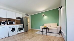 A compact living space with a kitchenette, a washing machine, a refrigerator, a microwave, and a small table with fruit.
