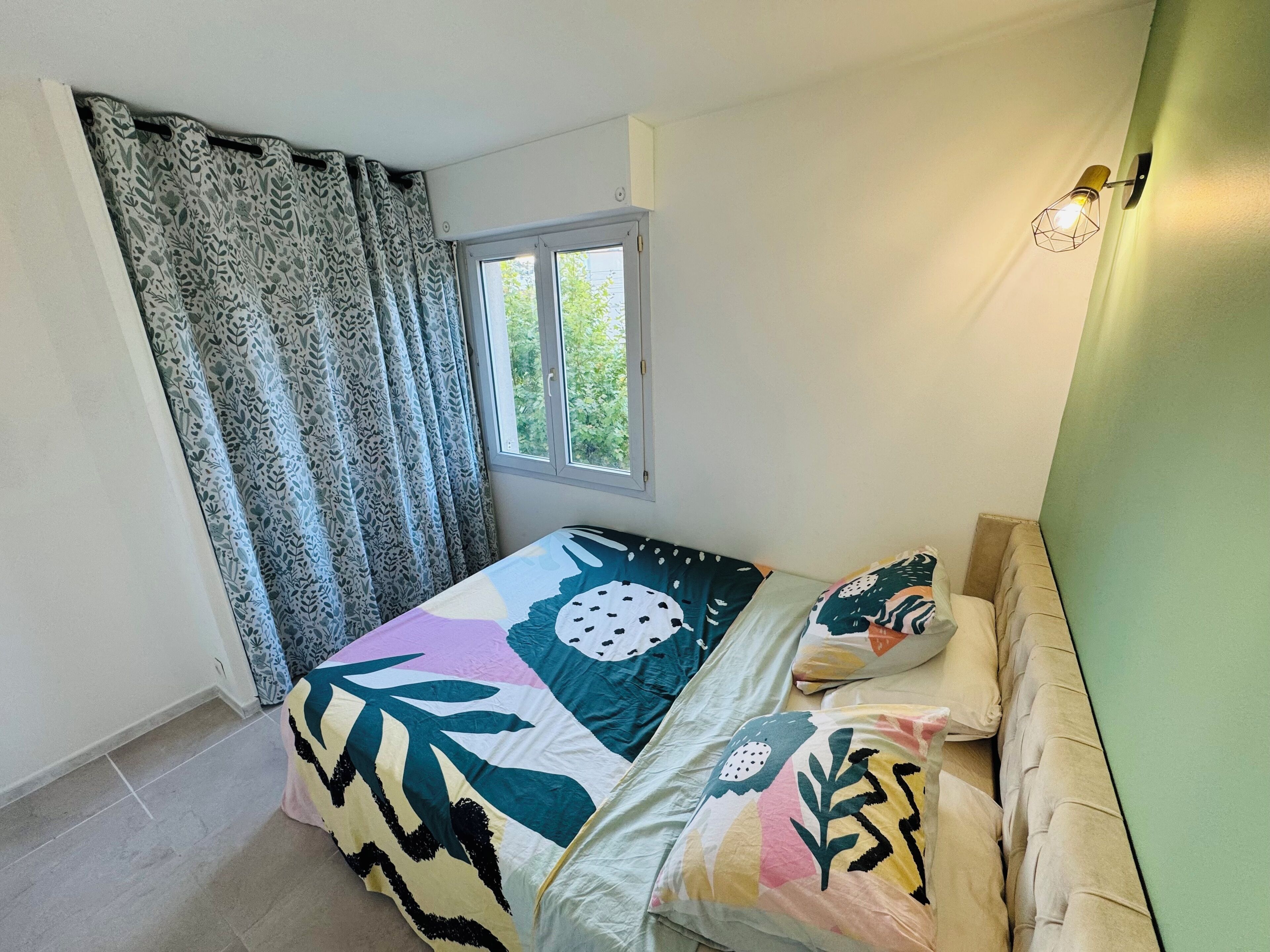 Un dormitorio con cama, cortinas estampadas, una ventana y una lámpara fijada a la pared.