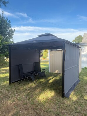 Een overdekt terras met stoelen en een tafel, naast een gebouw.