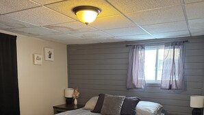 A bedroom with a bed, two bedside tables, a window with curtains, a ceiling light, and a wall-mounted picture.