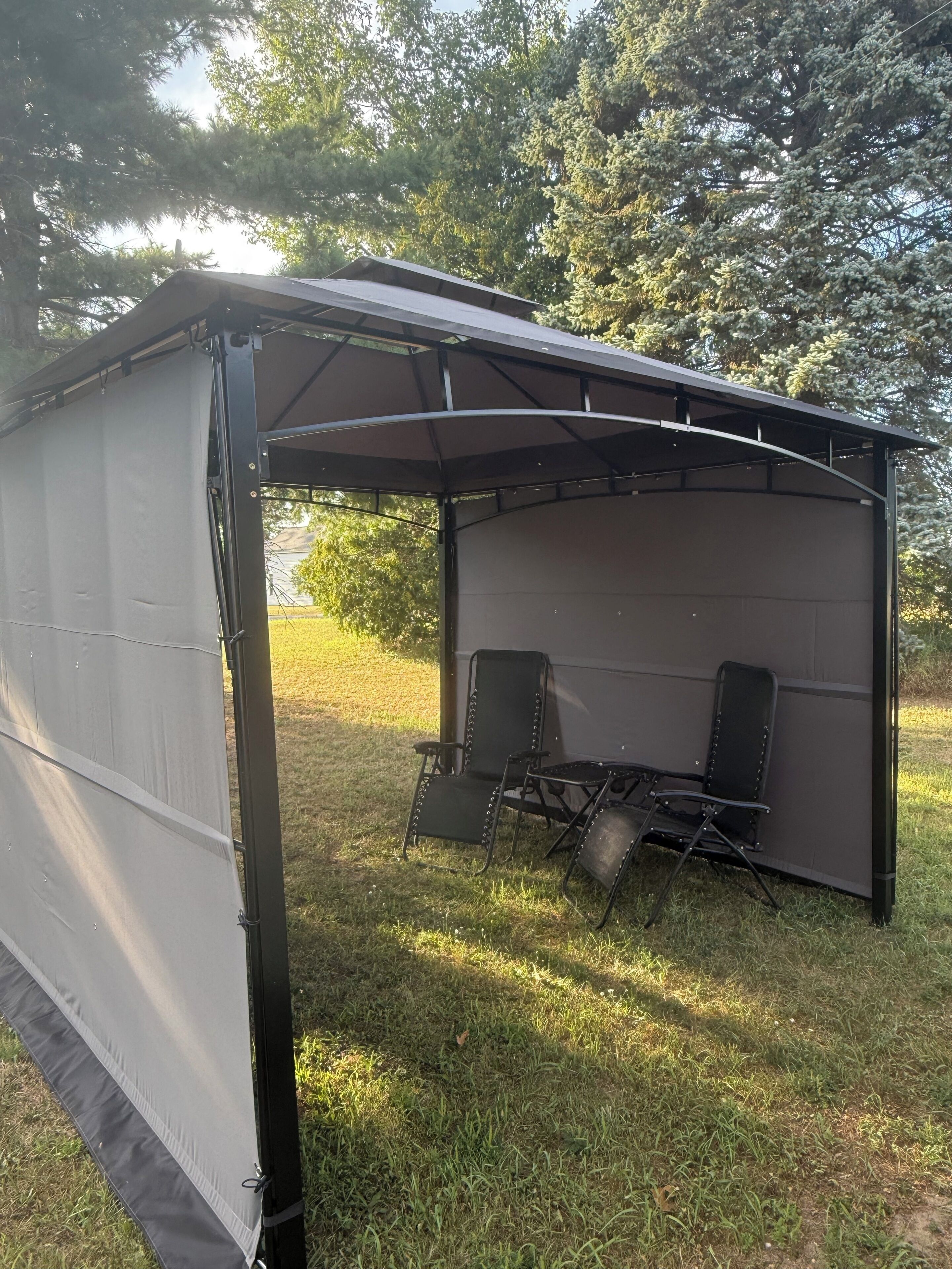 Un espace extérieur abrité, aménagé avec deux chaises et une table, entouré de pelouse et d’arbres.