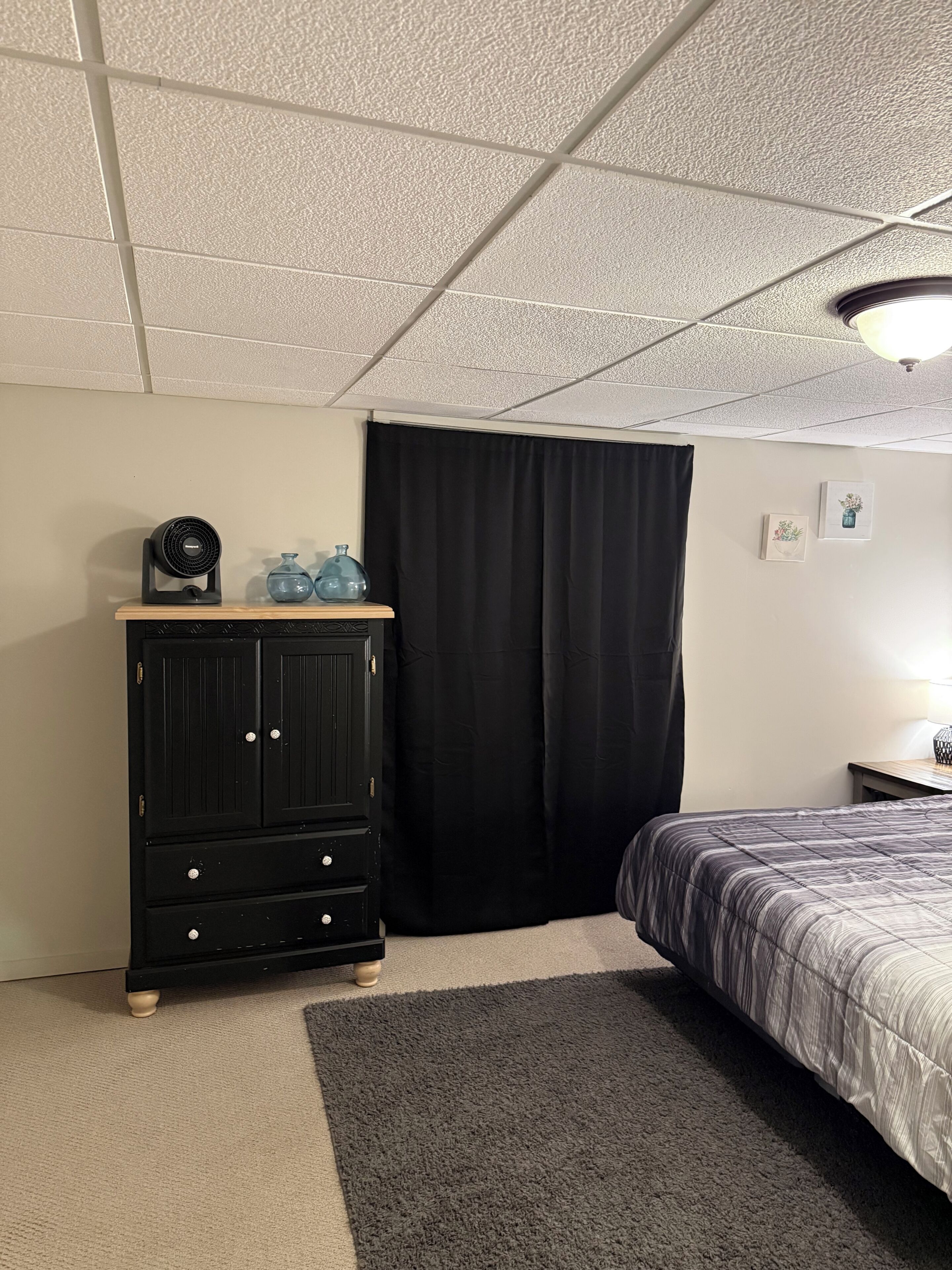 Une chambre à coucher avec un lit, une commode, un ventilateur et une fenêtre avec des rideaux.