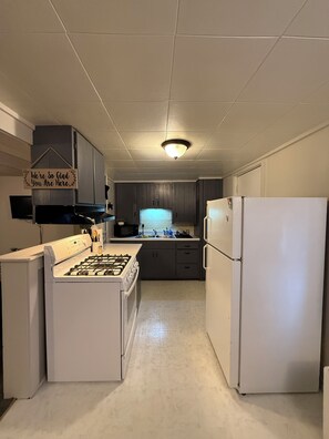 Een keuken met een gasfornuis, een koelkast en een magnetron. Er hangt een bord aan de muur en boven het aanrecht zijn kastjes.