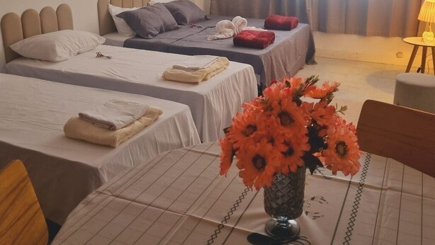 Habitación de hotel con dos camas, una mesa de comedor con un centro de mesa floral, un cuadro en la pared y un amplio ventanal con cortinas.