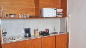 Et kompakt køkken med træskabe, en mikroovn, kaffemaskine og vask. Der står plantekasser på hylderne og en lille skraldespand ved siden af vasken.