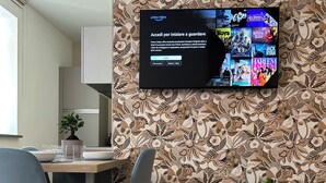 Un salon moderne avec un téléviseur à écran plat fixé sur un mur recouvert d’un papier peint à motifs floraux, un coin repas avec une table et des chaises en bois, et une plante en pot.