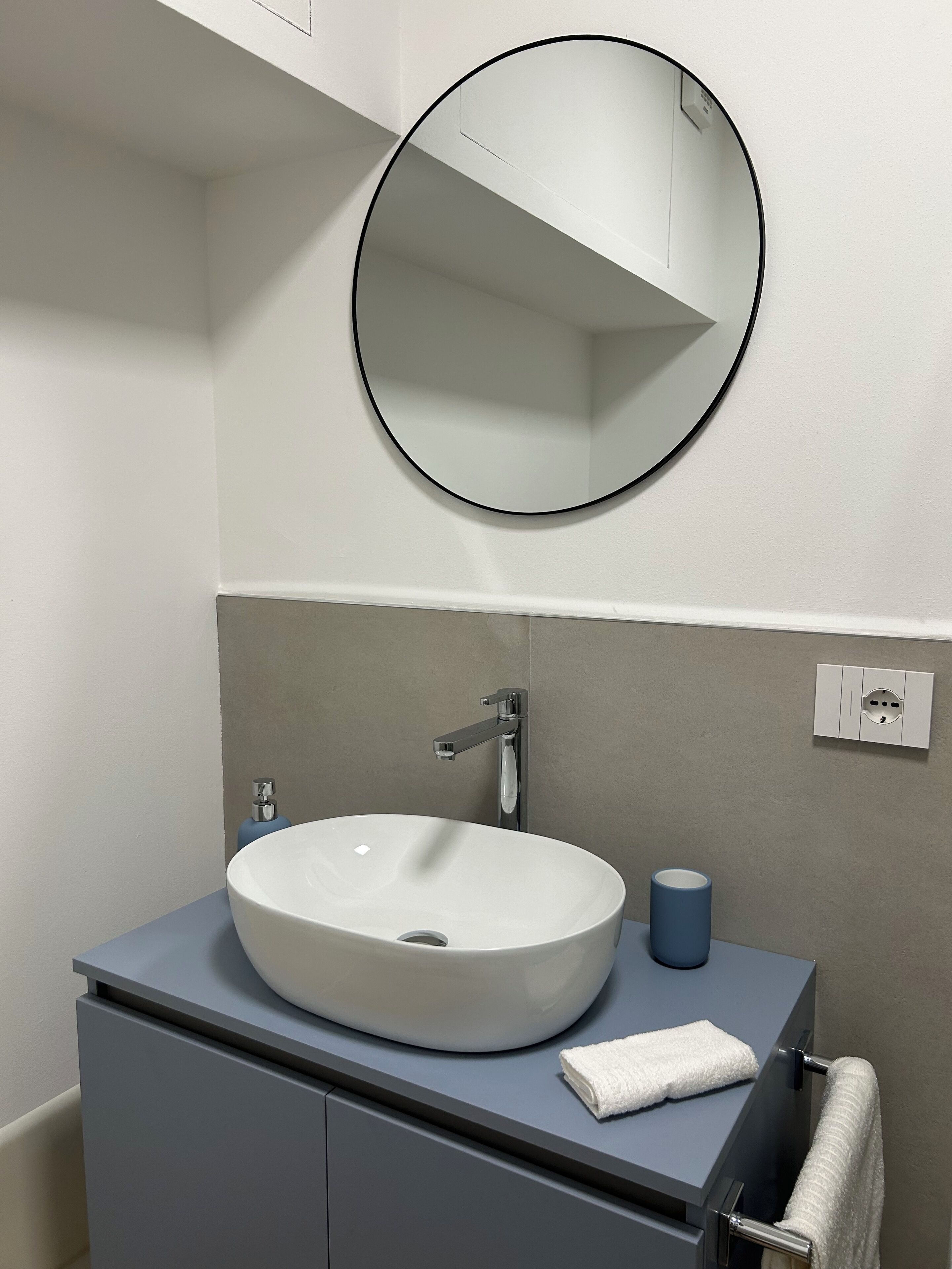 Un baño con un espejo redondo, un lavabo blanco, una toalla azul y un dispensador de papel higiénico.