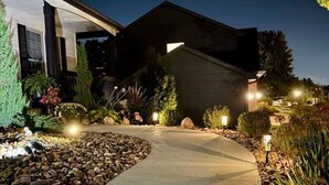 A well-lit pathway leading to a house with a dark roof, surrounded by landscaped gardens and decorative lighting. - Family-Friendly 5BR Home w/ Games & Massage Chair (Papillion)