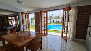 Un comedor amplio con mesa y sillas de madera, ventanas grandes con vista a una piscina y vegetación, además de una pared de ladrillo.