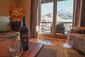 Uma garrafa de vinho Claret Francis Coppola e dois copos sobre uma mesa de madeira, num espaço com vista para uma montanha através de uma porta de vidro.