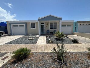 A modern two-story house with a garage, a front porch with chairs, and a landscaped front yard with various plants. - 3-bedroom house in beautiful Guanica with AC (guanica)