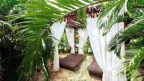 Un jardin tropical avec un hamac et un petit lit placé sous une pergola agrémentée de rideaux blancs.