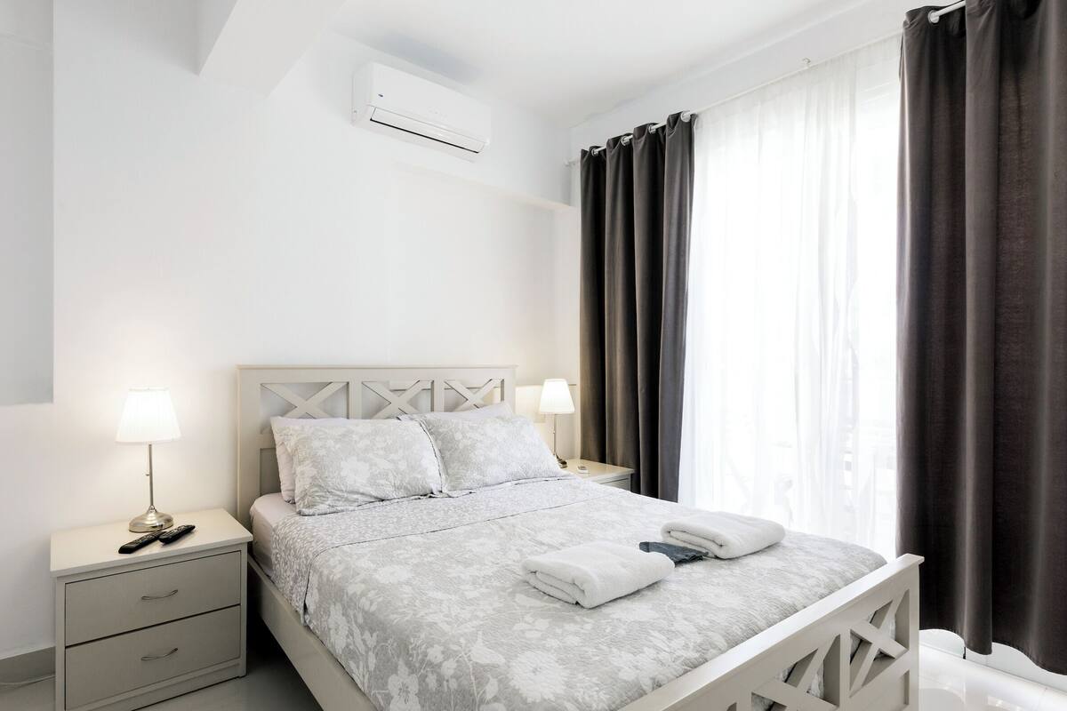 Une chambre à coucher avec un lit, des tables de chevet, une lampe, une télécommande et une fenêtre avec des rideaux.