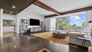 A modern living room with a large TV, wooden flooring, and a view of trees through large windows. - Sherman Oaks Oasis with Views and Pool Table (Los Angeles)