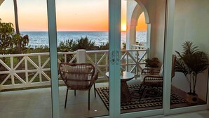 Ein Balkon mit Blick auf den Sonnenuntergang über dem Meer, mit Korbmöbeln, einem kleinen Tisch und einem Blumentopf.