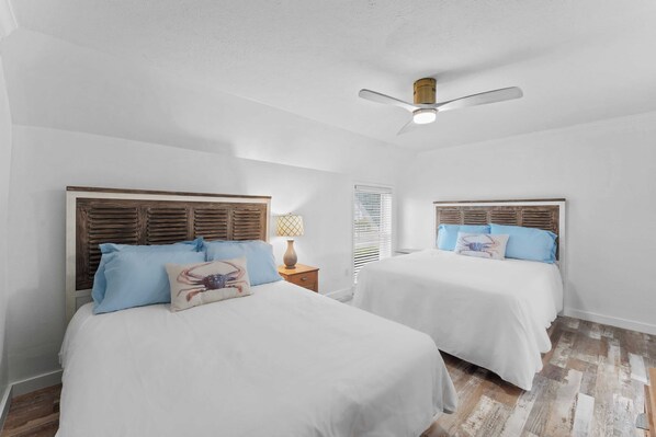 Une chambre avec deux lits, des têtes de lit en bois, du linge de lit blanc, un ventilateur de plafond et une fenêtre avec des stores.