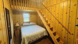 Une chambre avec un escalier en bois, un lit recouvert d’une courtepointe, une table de chevet avec une lampe et une fenêtre.