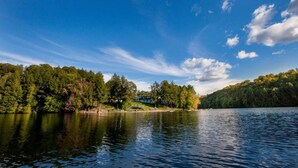 A serene lakeside view with dense forested hills, a clear sky, and calm water reflecting the surroundings.