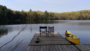 A wooden dock with two chairs and a canoe on a calm lake, surrounded by dense forest.