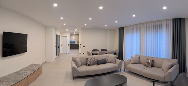 A modern living room with two sofas, a flat-screen TV, and a dining area in the background.