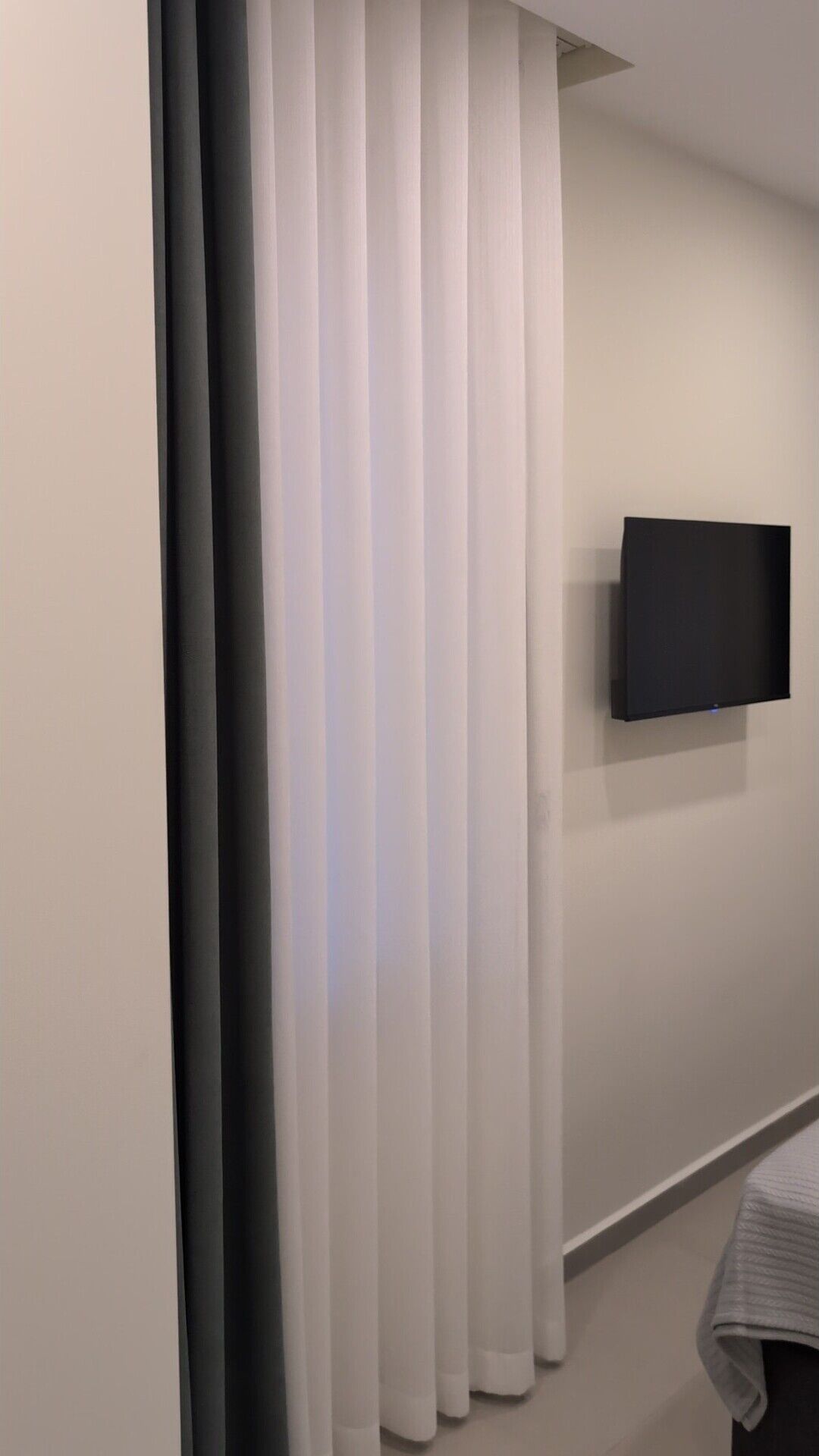 Una habitación con un ventanal grande cubierto por persianas verticales blancas y oscuras, un televisor de pantalla plana montado en la pared y una mesita con mantel blanco.