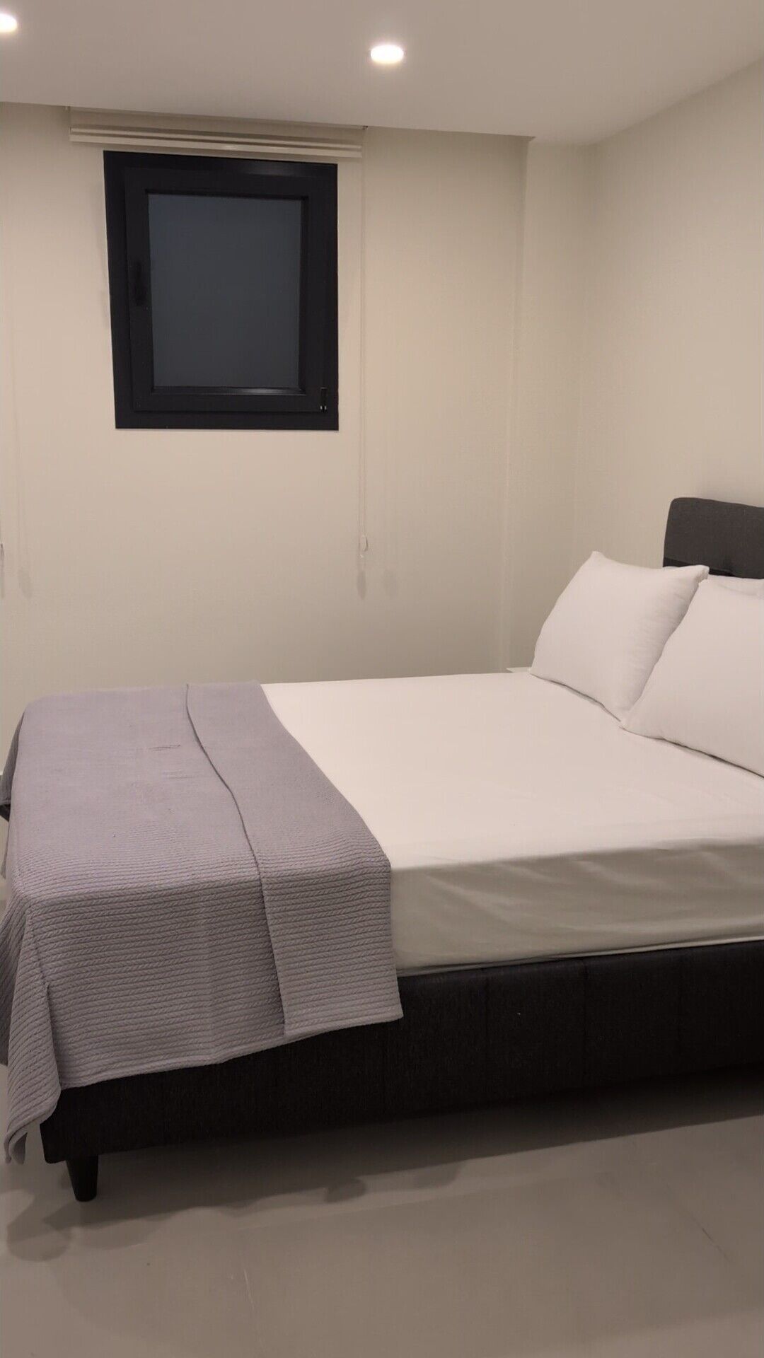 Una cama individual con una manta gris y almohadas blancas, cabecera negra y una ventana con marco negro.