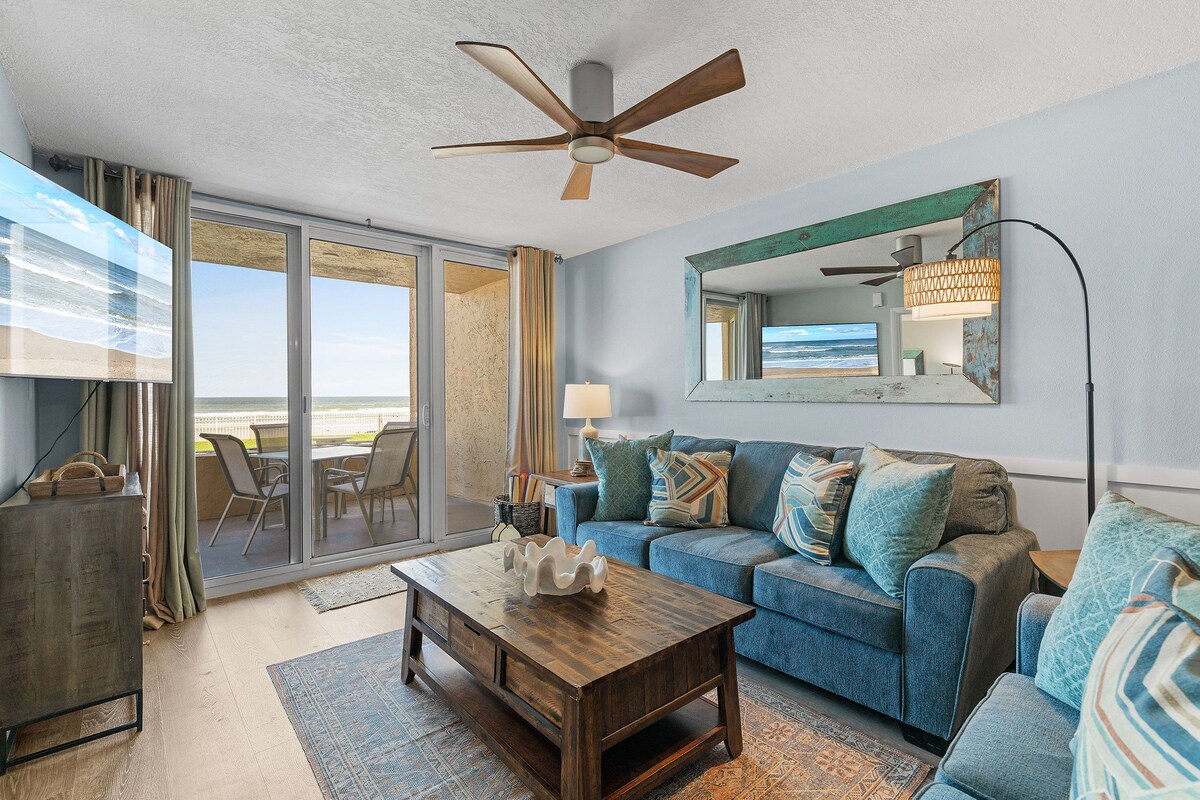 Un salon avec un canapé bleu, une table basse en bois, un ventilateur de plafond et une grande fenêtre donnant sur la plage.