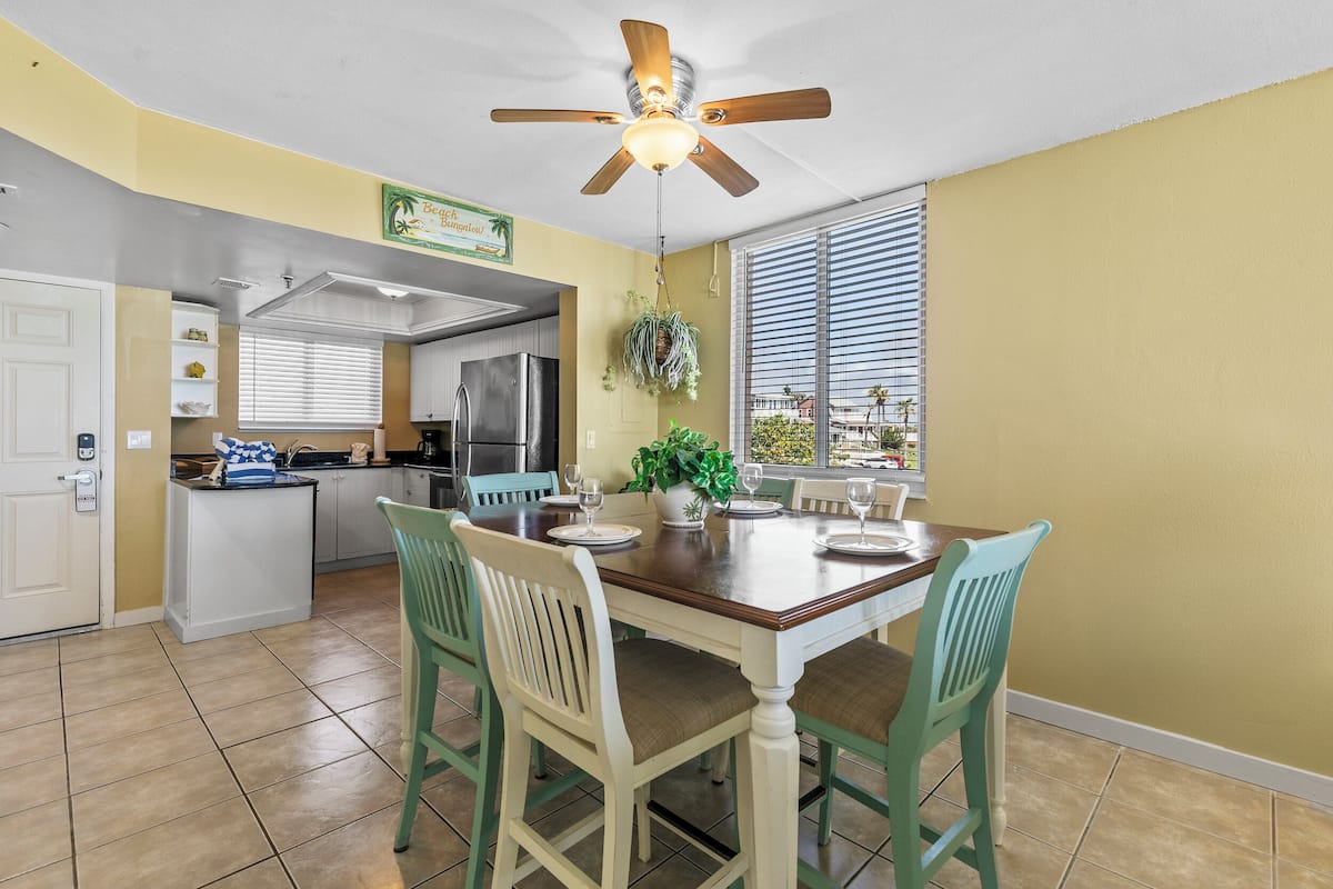 Une cuisine équipée d’une table à manger, de chaises, d’un ventilateur de plafond et d’une fenêtre avec des stores.