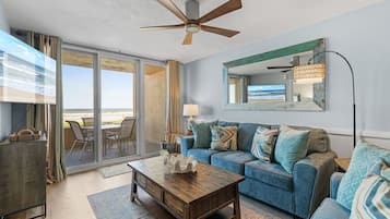 Un salon avec un canapé bleu, une table basse en bois, un ventilateur de plafond et une grande fenêtre donnant sur la plage.
